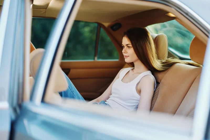 Young woman relaxing in the back seat of a car, reflecting on life with a calm expression, wearing a casual white tank top and denim jeans against a green blurred background.