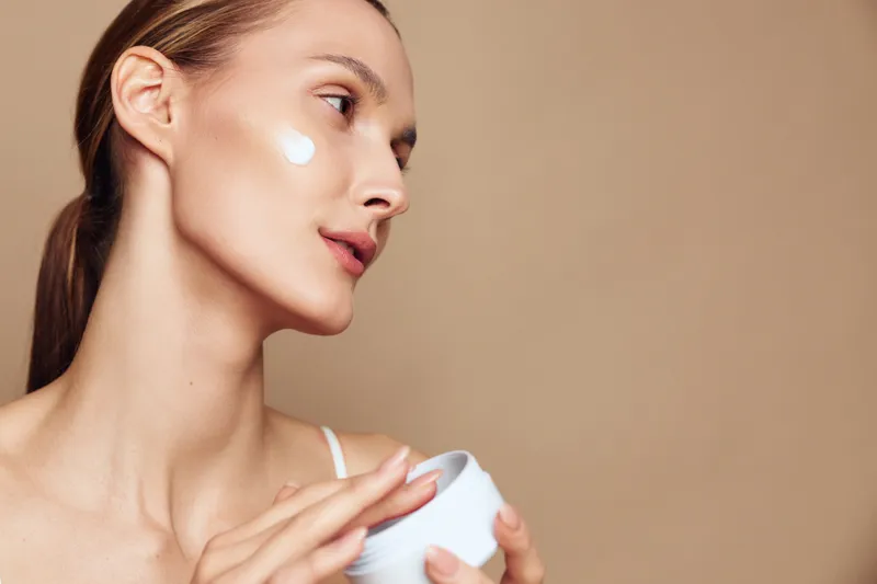 Young woman applies moisturizing cream on her cheek with a gentle touch, showing natural skin care routine and healthy complexion, isolated on beige background.