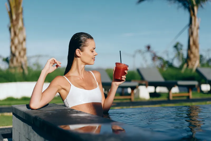 Poolside woman in a white bikini holds a refreshing iced drink, soaking up sunlight. Close-up lifestyle scene highlighting relaxation, wellness, self-care, and leisurely moments by water.