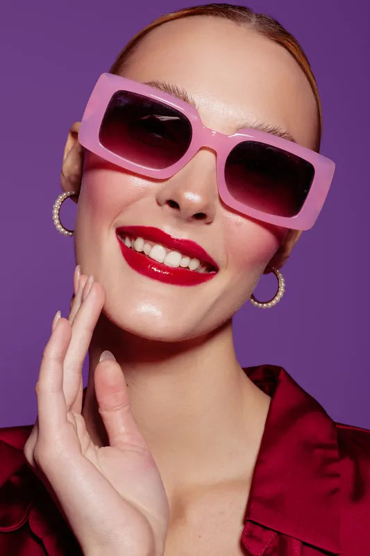Smiling woman with pink sunglasses and red lipstick poses against purple background. Fashion portrait with glossy skin, hoop earrings, and confident expression in studio lighting.