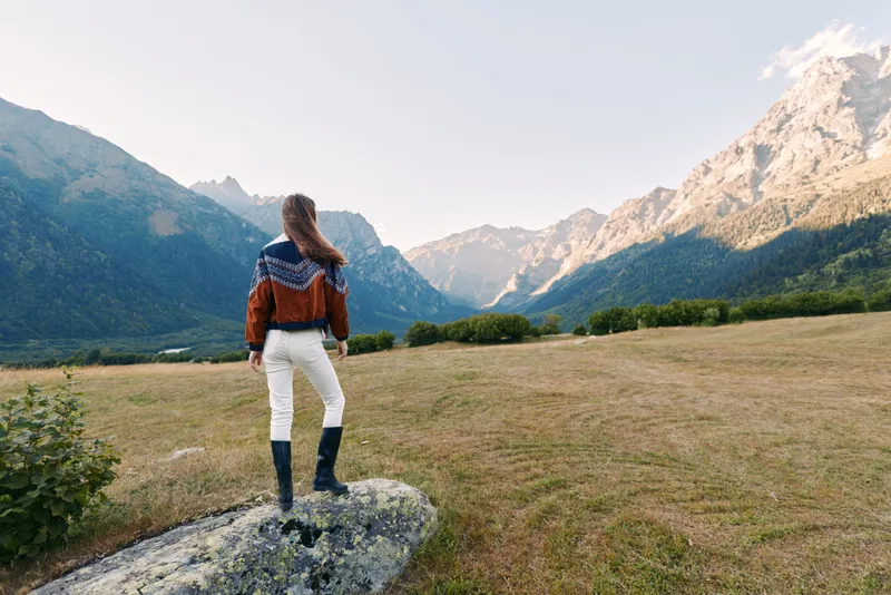 Woman stands on rock overlooking mountains landscape and nature meadow, hiking outdoors enjoying panoramic view at sunrise. Traveler in sweater and boots exploring alpine valley.