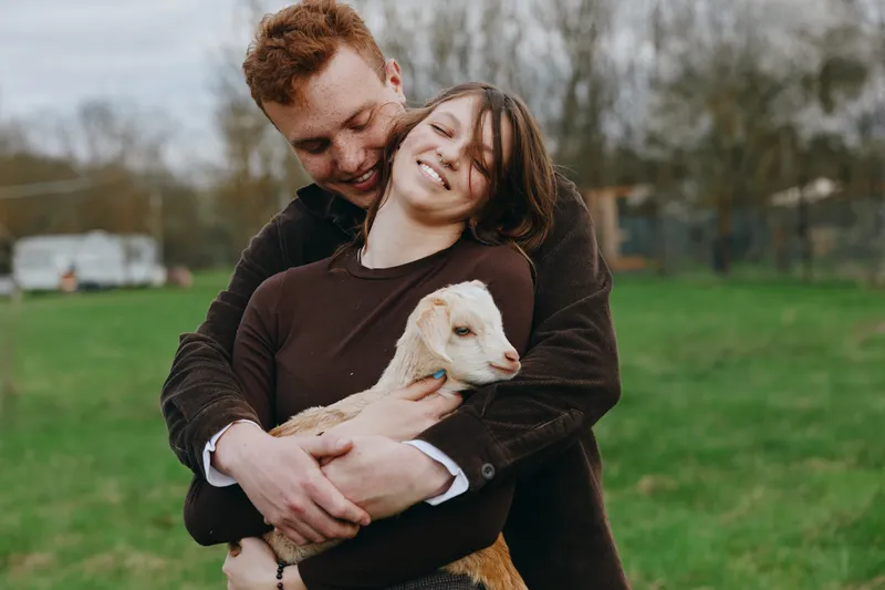 Smiling couple embraces a young goat kid in a sunny field, showcasing countryside lifestyle and bonding through animals. Tender moment under soft natural light evokes warmth and happiness.