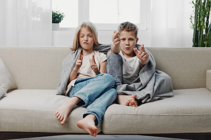 Two playful children sit barefoot on a beige sofa making funny faces and hand gestures in a bright living room. Concept of childhood fun, creativity, and sibling bonding.