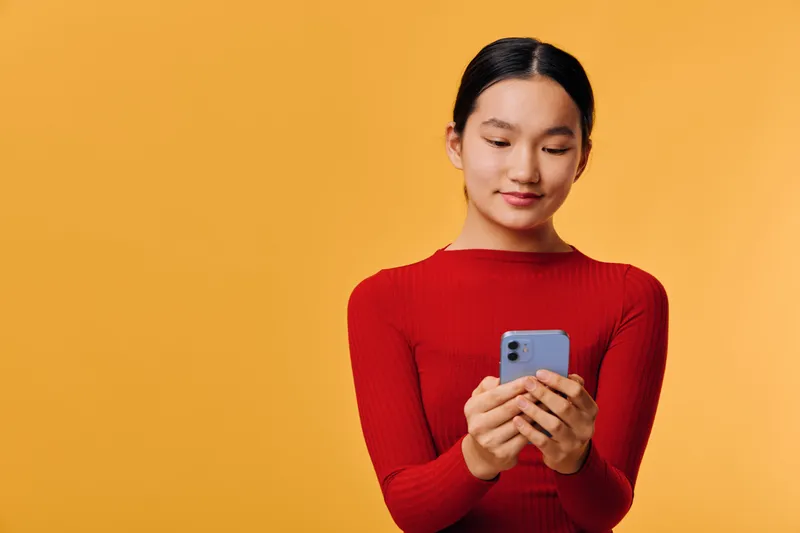 Young woman in red sweater holding smartphone, looking at screen with slight smile on yellow background. Technology, communication, and casual lifestyle concept with simple colorful backdrop.