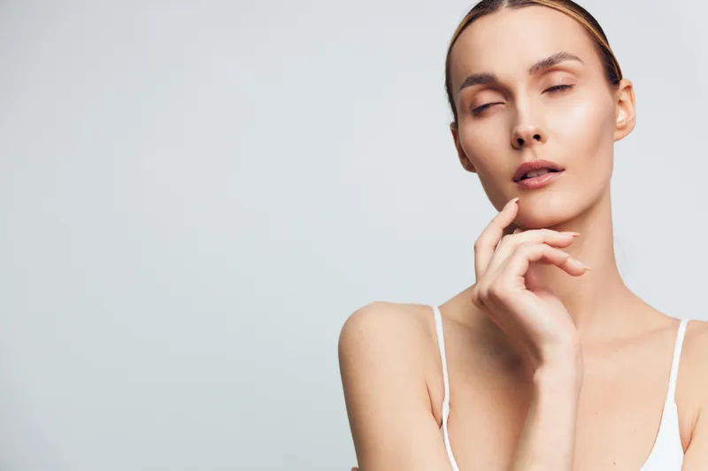 A young woman with clear skin poses with closed eyes and a relaxed expression, wearing a white tank top against a plain light background. Concept for beauty and skincare.