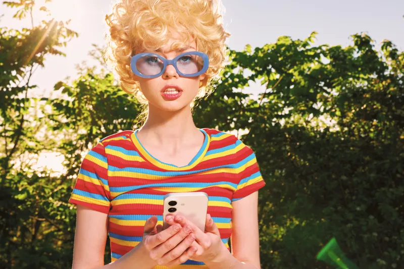 Young woman with curly hair wearing a bright striped t shirt stands outdoors holding a smartphone, eyes fixed on the screen, a playful grin spreading across her face