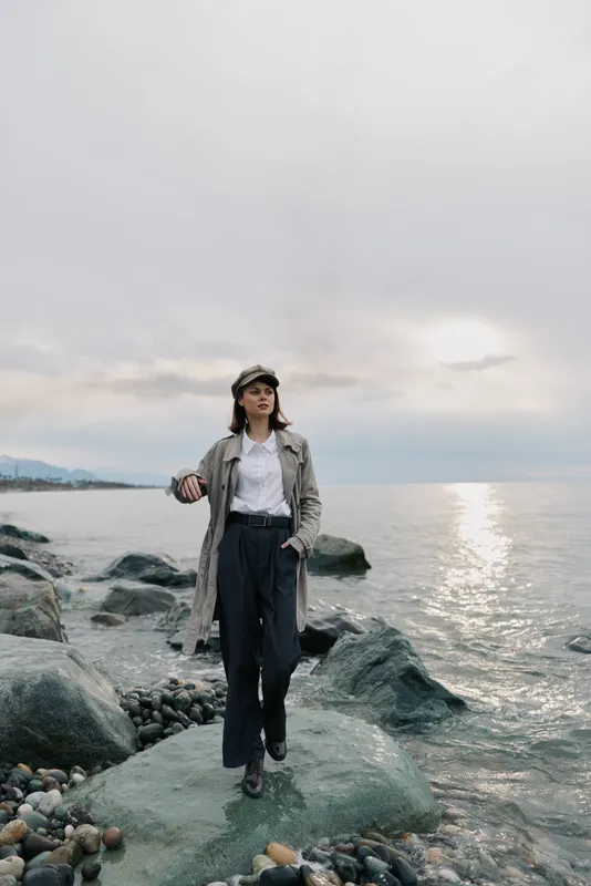 Young woman walks on rocky shore during cloudy sunset wearing trench coat and wide pants. Concept of fashion, outdoor lifestyle, and peaceful coastal scenery.