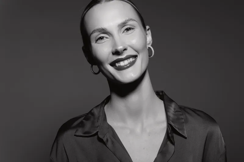 Portrait of a smiling caucasian woman with fair skin and slicked back hair wearing hoop earrings and a dark blouse. Studio lighting creates a confident and elegant mood.