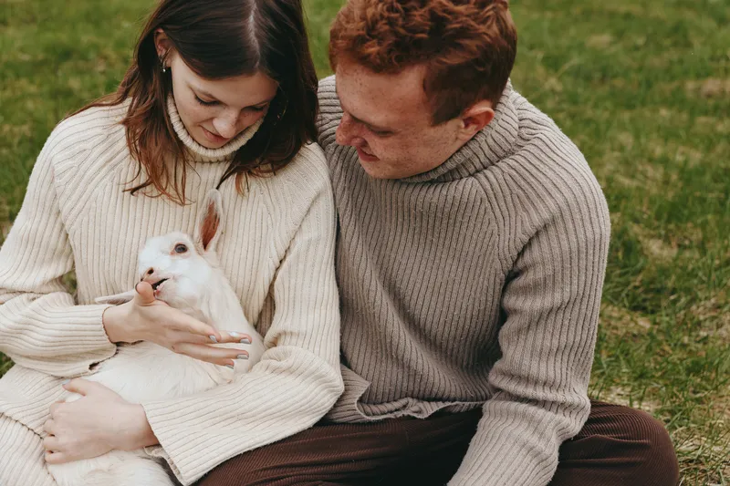 Relaxed couple in cozy sweaters holds a white rabbit in a sunny outdoor setting, capturing a warm lifestyle moment of affection, companionship, and care with soft lighting and gentle colors.