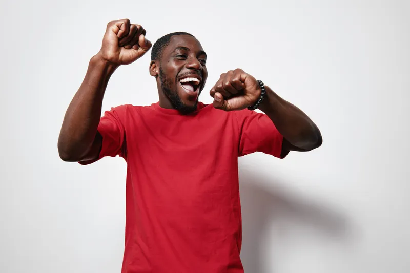 African American man with dark skin wearing red t shirt dancing and smiling with raised hands on white background. Happy young black male enjoying music and movement.