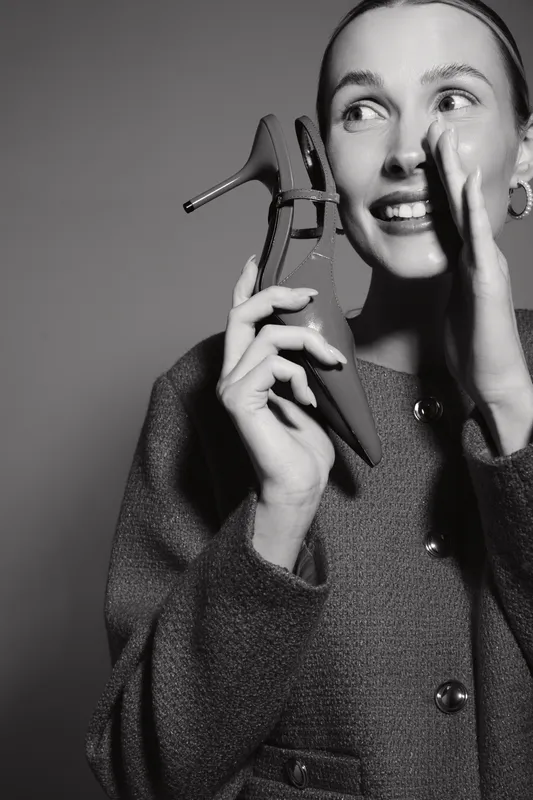 Smiling Caucasian woman with light skin holding high heel shoe near her face wearing coat. Concept of fashion, happiness, and style in studio lighting.