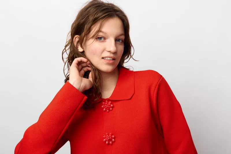 Young woman with curly hair wearing red sweater poses with hand near face against white background. Casual style, natural look, and confident expression in studio portrait.