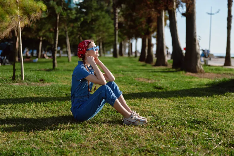 thoughtful woman sitting on grass wearing blue outfit and red headband in sunny park with tall trees and walkway background