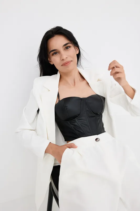 Young woman wearing a black corset and white blazer posing confidently against a plain white background with a stylish, modern look in a studio setting.