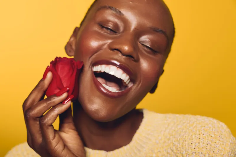 Beautiful dark skinned African woman with closed eyes smiling widely holds red rose near face on yellow background. Concept of happiness, beauty, love, and natural skin care.