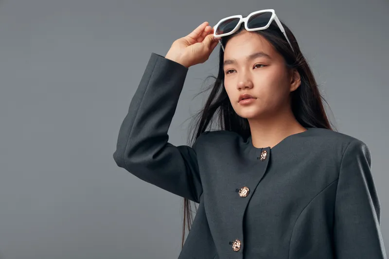 Young woman adjusting white sunglasses with hand while looking away in dark stylish jacket against neutral background. Portrait of fashion model with confident expression and long hair.