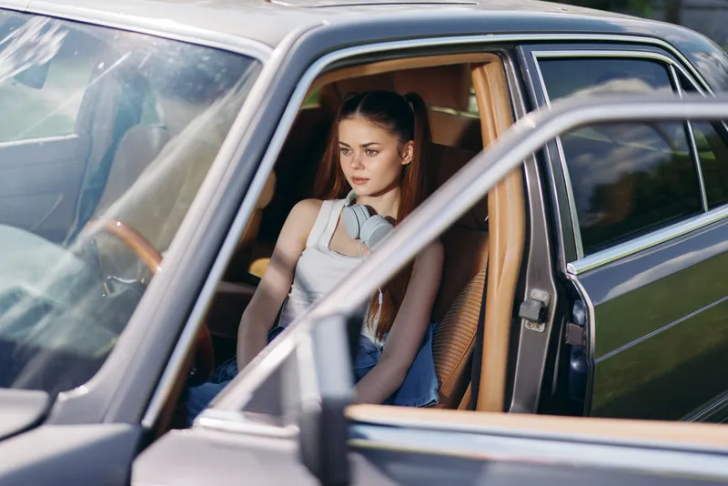 Young woman with headphones, sitting in a vintage car, enjoying music outdoors on a sunny day, relaxed mood, looking contemplative.