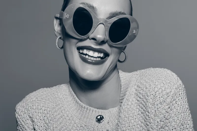 Happy smiling caucasian woman with light skin wearing sunglasses and knitted sweater poses against gray background. Concept for fashion, lifestyle, and positive emotions.