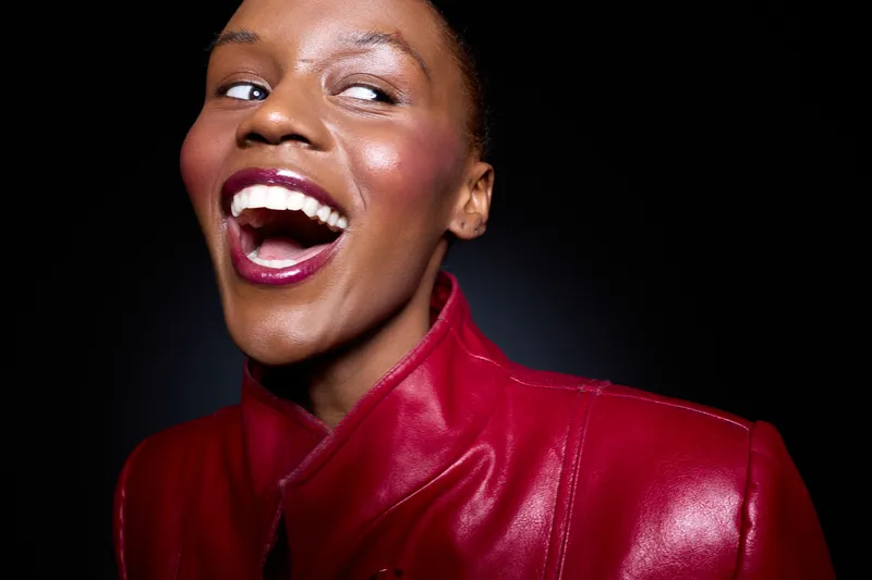 Happy African American woman with dark skin laughing, wearing a bright red leather jacket. Studio portrait on black background. Concept of joy, confidence, and fashion.