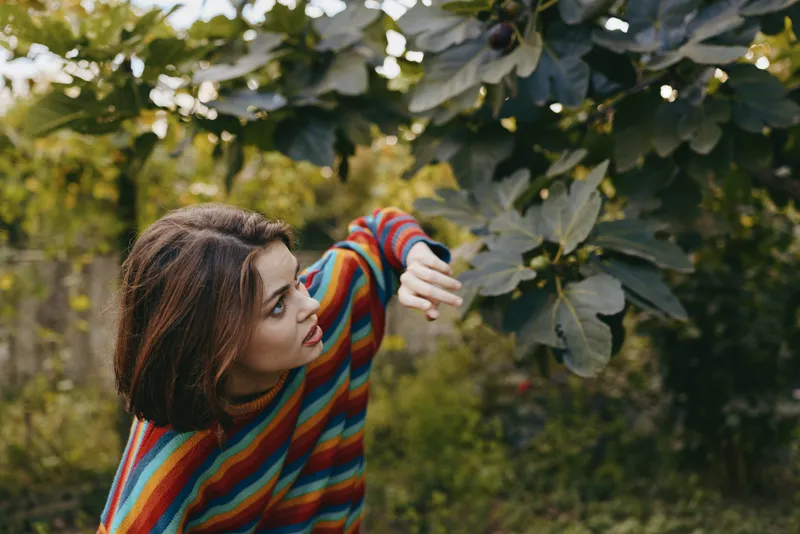 Young adult woman in a colorful striped sweater reaching for large leaves in a garden, curious and contemplative outdoor portrait with natural light and green foliage.