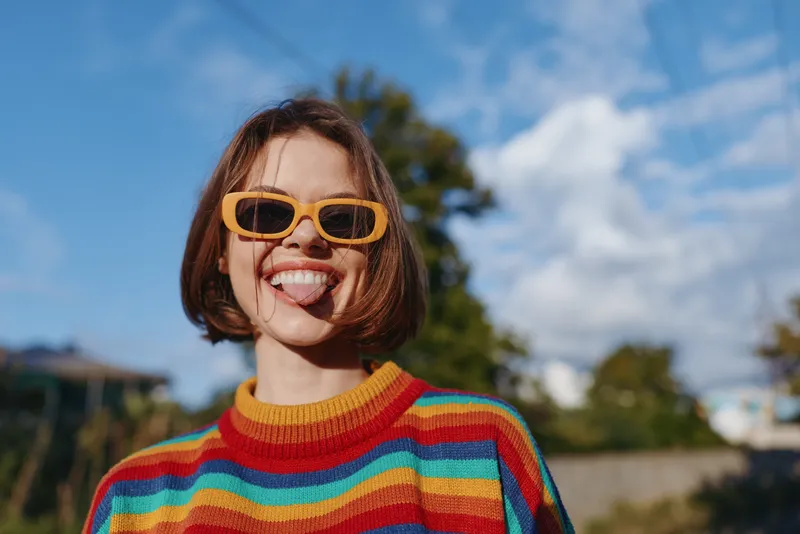 Woman in rainbow sweater and yellow sunglasses, playful and smiling while sticking tongue out. Young adult with short hair in casual outfit outdoors, vibrant fashion lifestyle portrait.