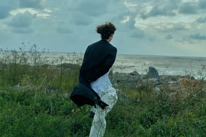 Windy coastline scene with a figure in a dark jacket running along grass by the sea, cloudy sky above, capturing motion and freedom in a dramatic coastal landscape