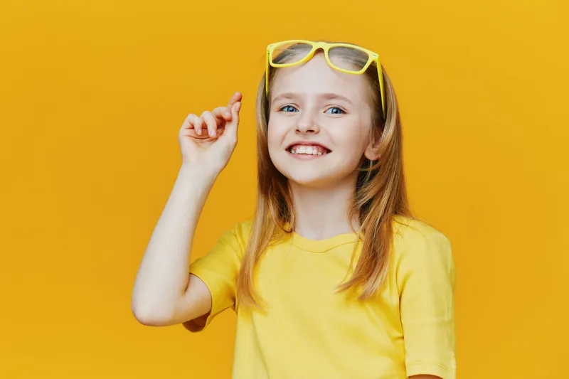 Cheerful girl with long hair wearing yellow sunglasses and a bright yellow t shirt, smiling and gesturing playfully against a solid yellow background