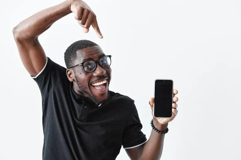 African American man with dark skin and glasses smiling and pointing at smartphone screen in black polo shirt on white background. Happy male showing mobile phone with excitement.