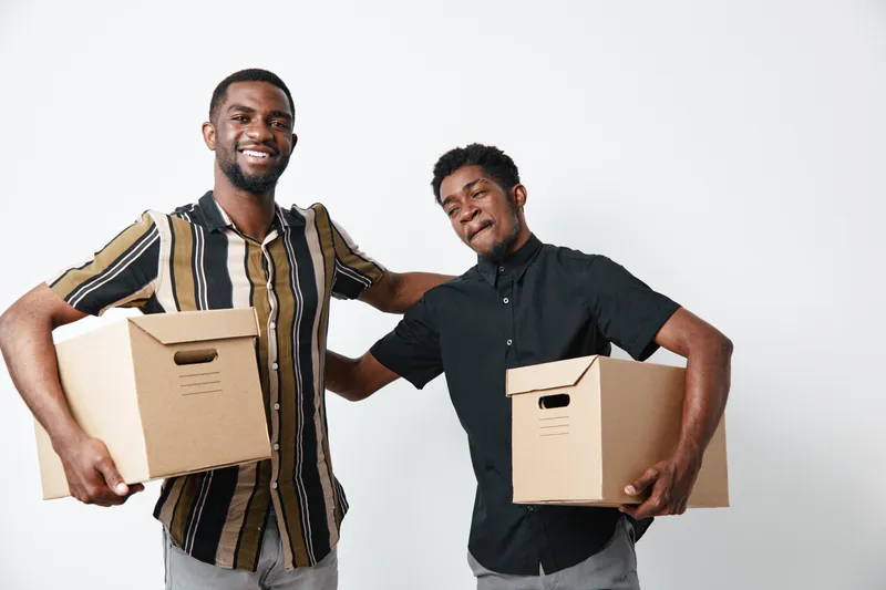 Two young black men holding cardboard boxes for moving or delivery, smiling and standing close together on white background, casual clothing and friendship.