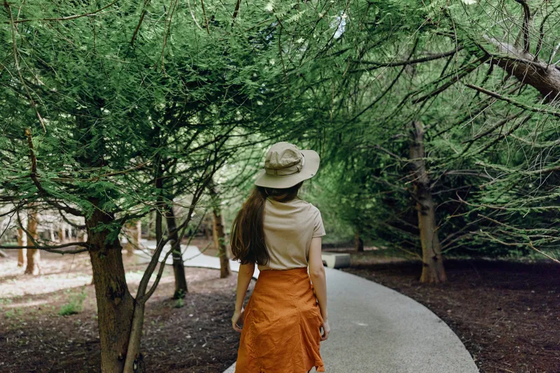 woman walking path trees hat back park skirt in green canopy, rear view of a traveler on a curved walkway surrounded by foliage for nature stroll and summer leisure