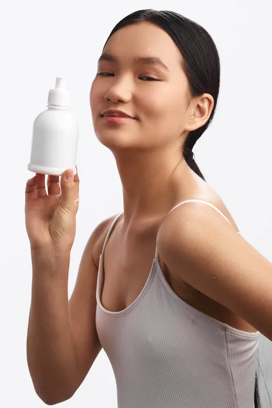 Asian woman in a clean studio holds a skincare bottle near her face, smiling softly as she showcases the product, conveying confidence, calm, and wellness in a modern lifestyle setting.