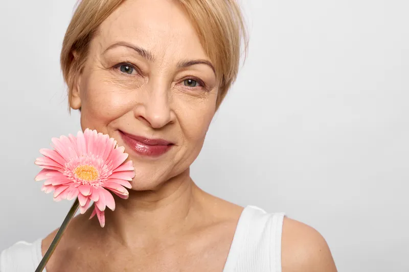 Smiling mature woman, closeup portrait, holding pink flower, gentle skincare, wellness mood, soft studio light, clean white background, selfcare, calm look