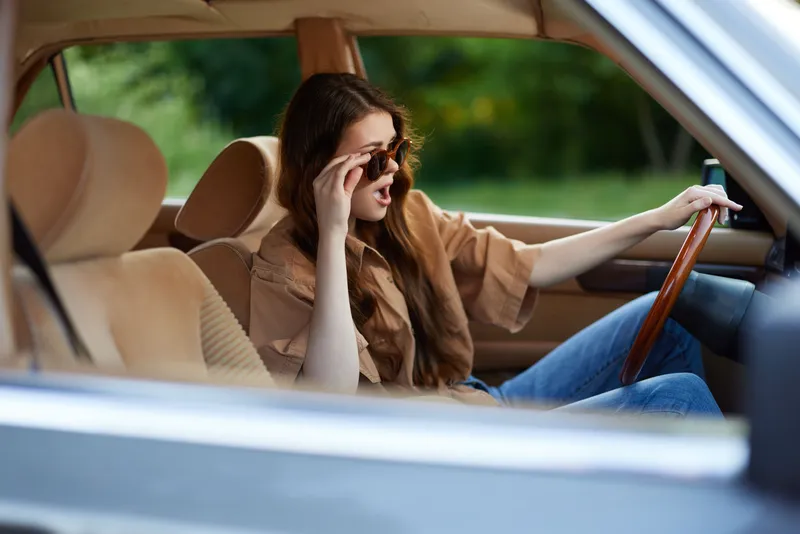 Stylish young woman driving a classic car with sunglasses, exuding confidence and joy in a beautiful outdoor setting.