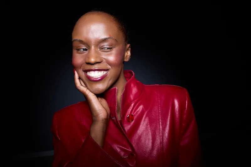 Smiling African woman with dark skin wearing a red leather jacket poses confidently on a black background. Concept of fashion, beauty, and empowerment for dark skin models.