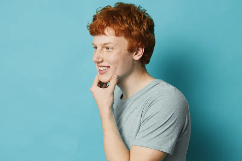 Young man with red hair and casual gray t shirt smiling thoughtful with hand on chin on blue background. Profile view, emotion and positive facial expression for lifestyle portrait.