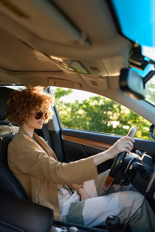 A woman with curly hair wearing a tan coat drives a car, wearing sunglasses, hands on the wheel as sunlight streams through the window, presenting a confident solo driving lifestyle.