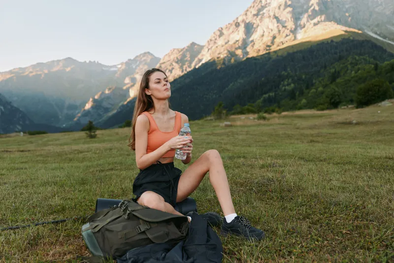 A relaxed woman sits on a grassy meadow in a mountain landscape, sipping water from a bottle. She wears athletic clothing and looks calm, enjoying a peaceful outdoor moment.