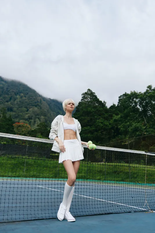 Stylish young female athlete in white sportswear poses confidently on a tennis court, showcasing fashion, sport, and summer vibes amidst a green backdrop