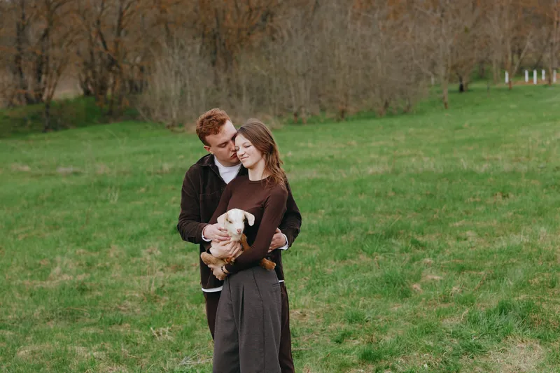 Young couple embraces tenderly in a grassy field while the woman holds a small baby goat, sharing a quiet and affectionate moment in casual autumn clothing.