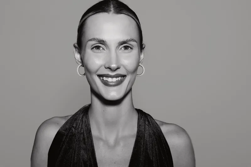Smiling caucasian woman with light skin and slicked back hair poses wearing sleeveless black top and hoop earrings in studio. Concept of beauty, fashion, and confident female model.