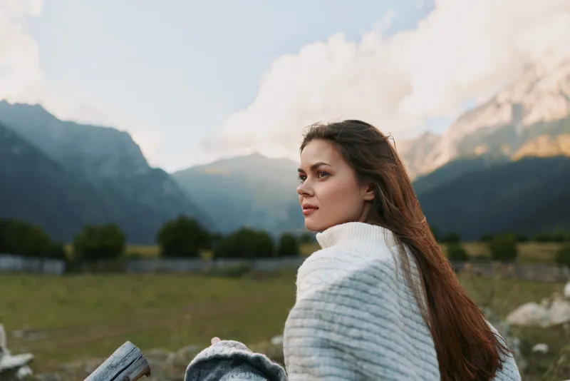 Young woman with long brunette hair gazes over a mountainous landscape in a white knit sweater, standing among grassy fields as distant peaks rise under a clear blue sky