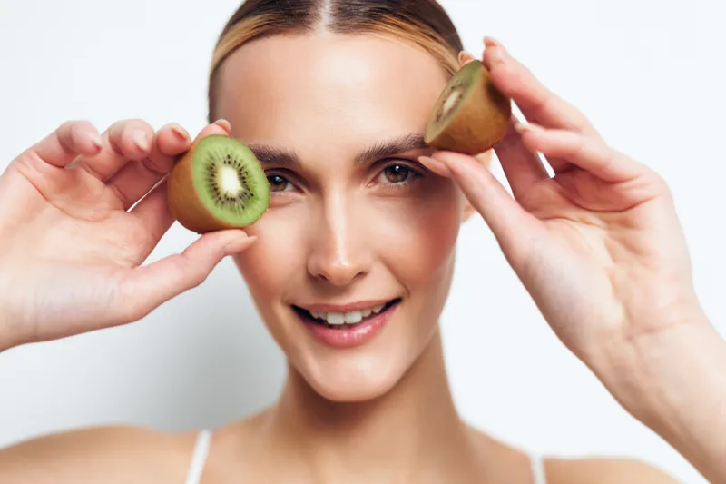 Young woman with blonde hair holds two halves of a kiwi fruit near her face and smiles softly against a white background.