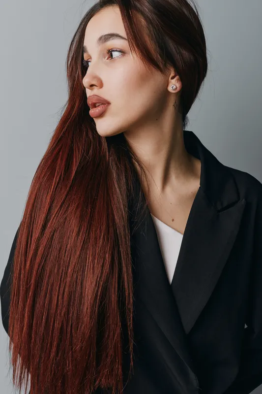 Young woman with long straight hair and natural makeup wearing black coat looking sideways on gray background. Portrait of female model with earrings and serious expression.