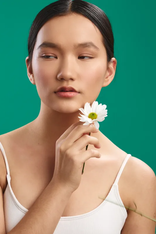 asian woman with a delicate face holds a white daisy against a vibrant green background, creating a calm and graceful portrait that highlights natural beauty and gentle mood.