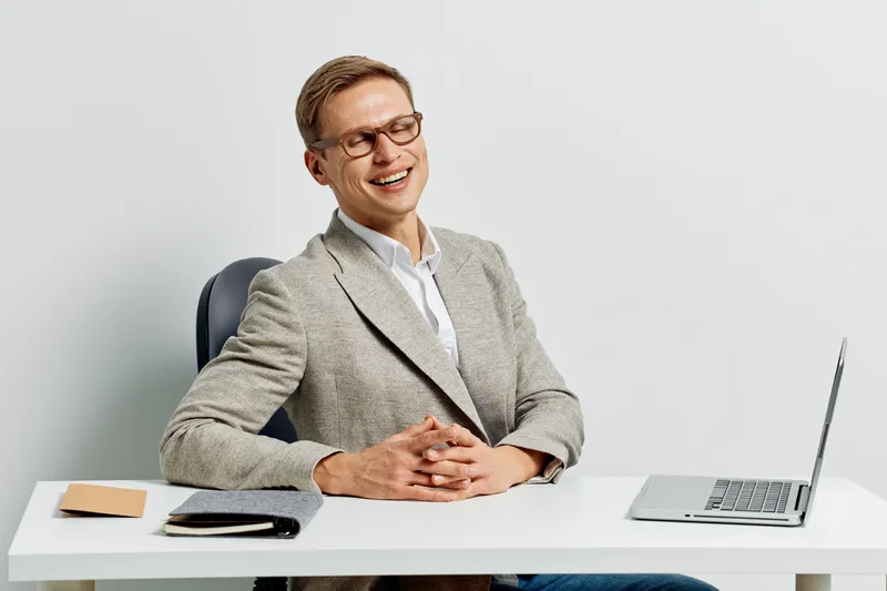 Smiling professional man in glasses and gray blazer sitting at desk with laptop in modern office background, demonstrating confidence and engagement in work, suitable for business and workplace concep