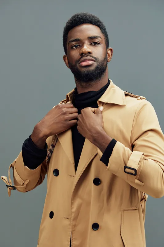 Stylish young man in a beige trench coat exuding confidence and charm against a neutral gray background, perfect for fashion and lifestyle concepts