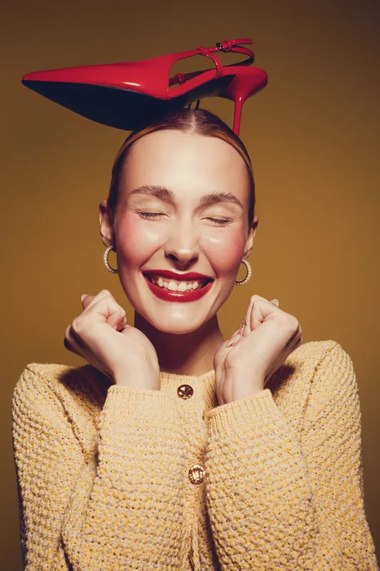 Happy woman with red lipstick and closed eyes smiles joyfully wearing a yellow sweater and balancing a red high heel shoe on her head. Concept of fun, fashion, and playful mood.
