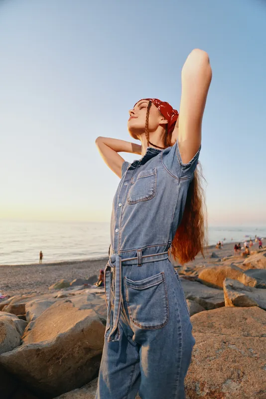 Lifestyle woman in modern boho-western denim street style poses confidently by the rocky beach at sunset with warm film color tones enhancing the natural mood.