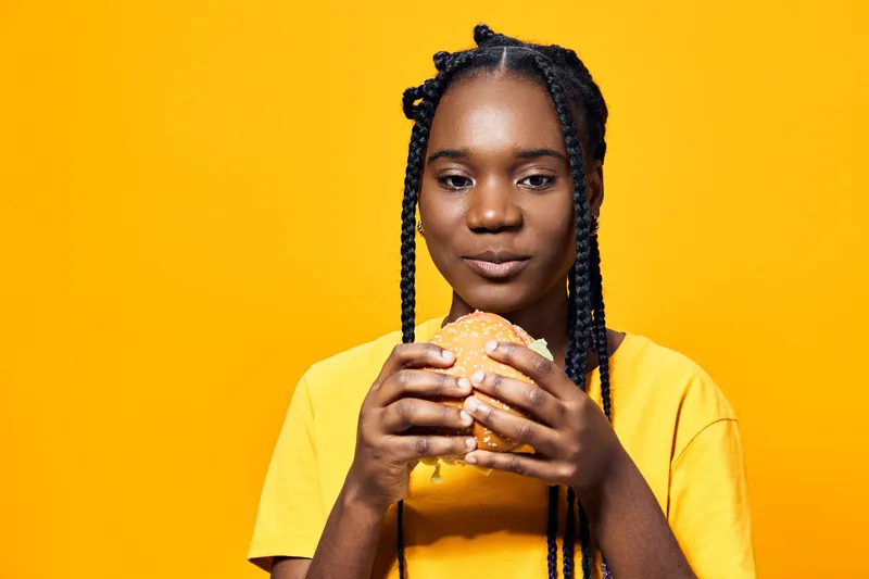 Portrait of a young African girl with braided hair enjoying a delicious hamburger against a vibrant yellow background, showcasing healthy eating concepts