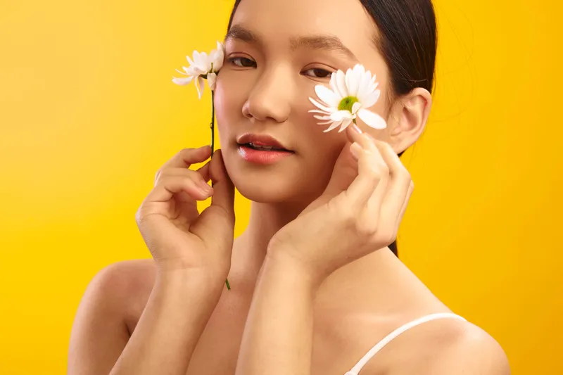 Asian woman with delicate features holds white flowers against a vibrant yellow backdrop, creating a fresh, minimalist portrait that emphasizes natural beauty and calm, sunny mood.